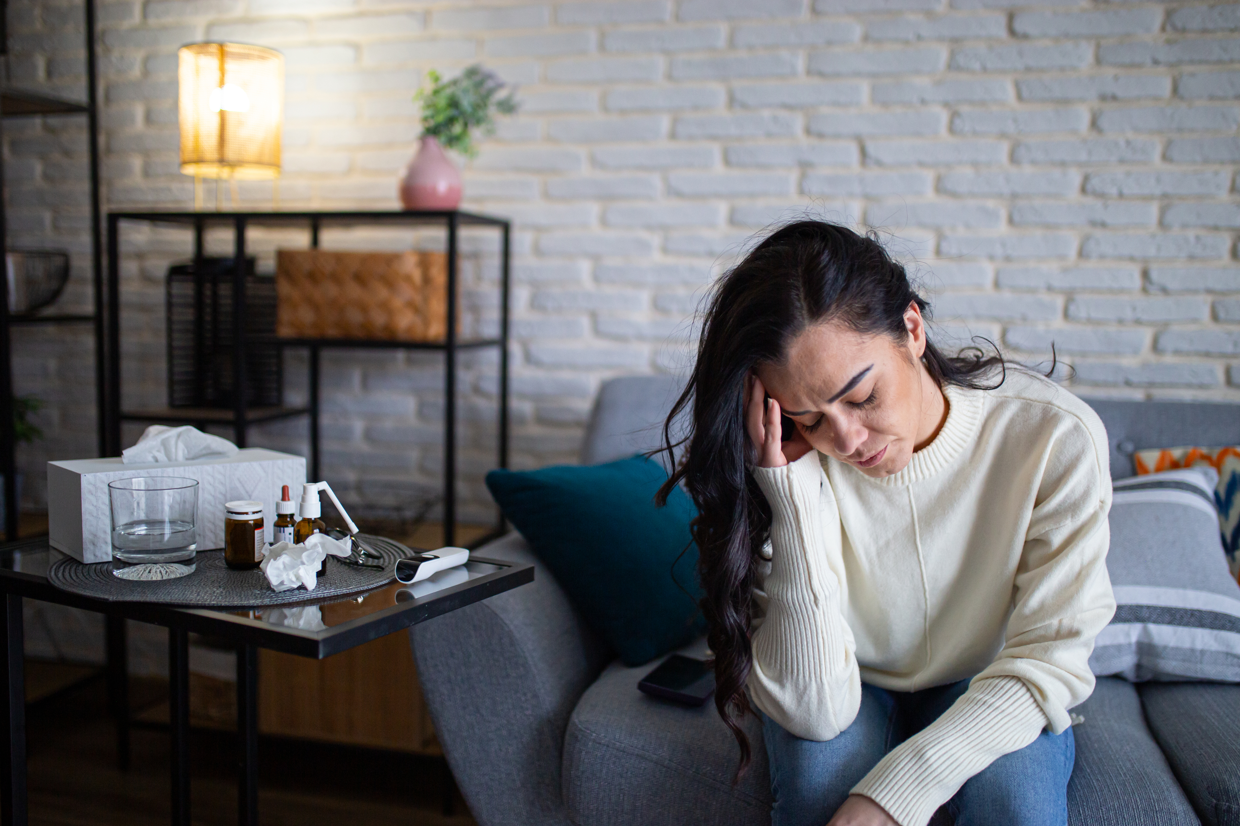 Kvinne i hvit genser som sitter på sofaen, ser uvel ut; Vev og medisin på sidebordet