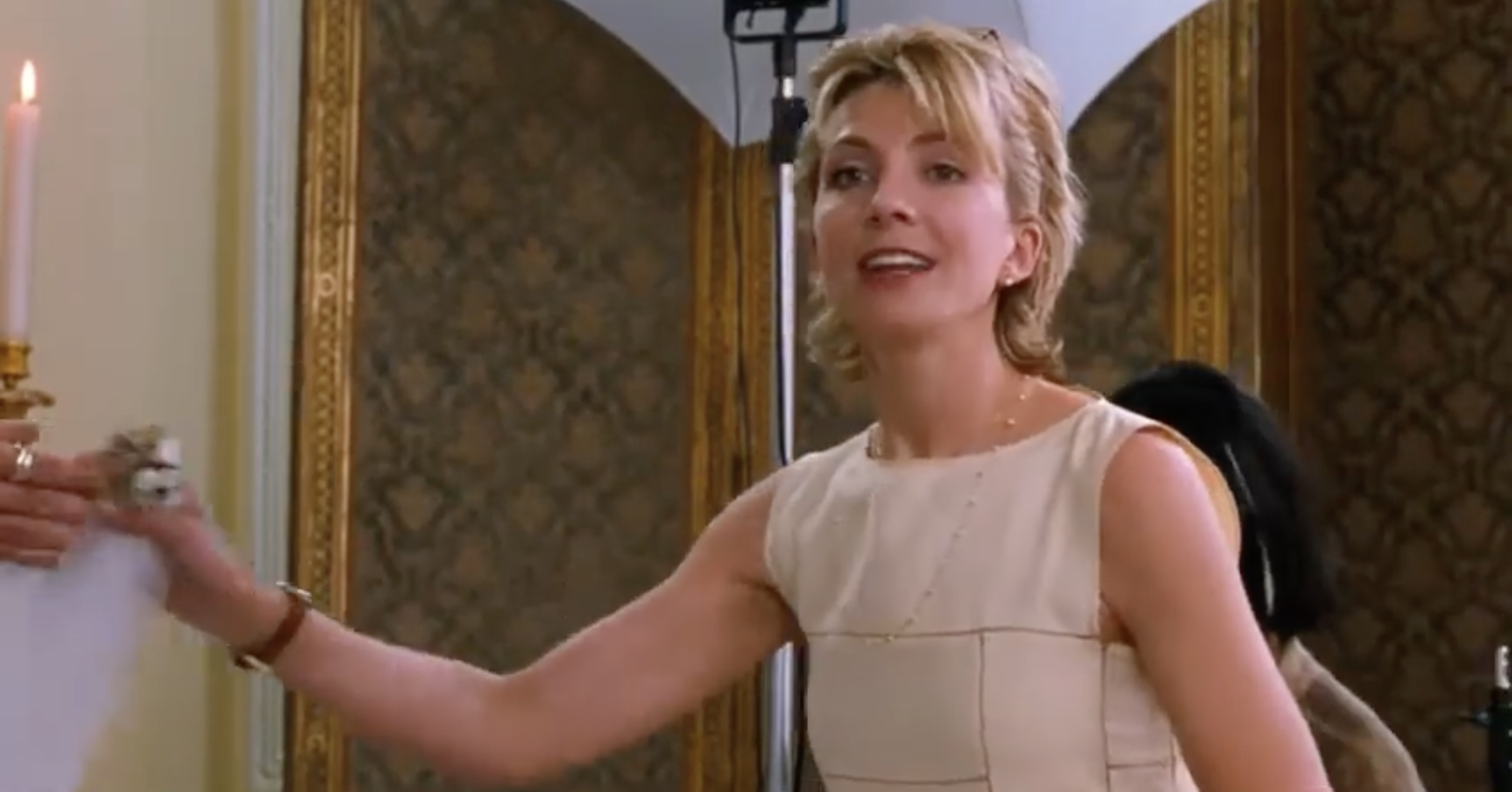 Person smiling, wearing a sleeveless dress, reaching out to shake someone's hand. Elegant setting with candle and patterned wallpaper in background