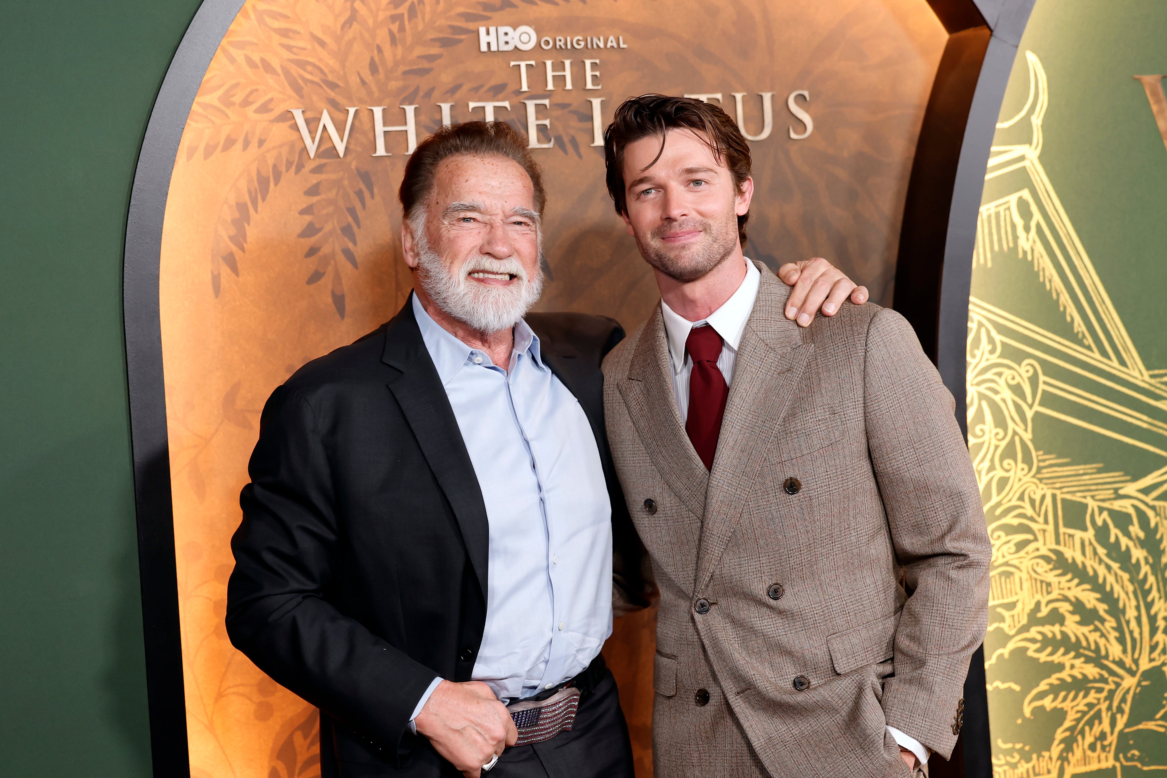 Two men pose together at an event for "The White Lotus." One wears a classic suit, the other a casual open-collar shirt and jacket