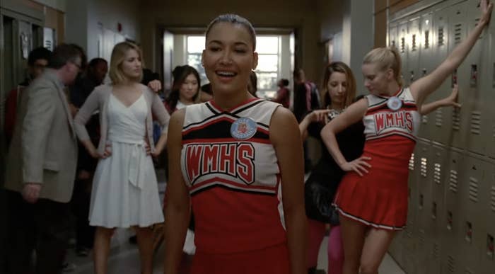 Group of cheerleaders in a school hallway, with one cheerleader smiling in front. Others stand or lean against lockers in the background