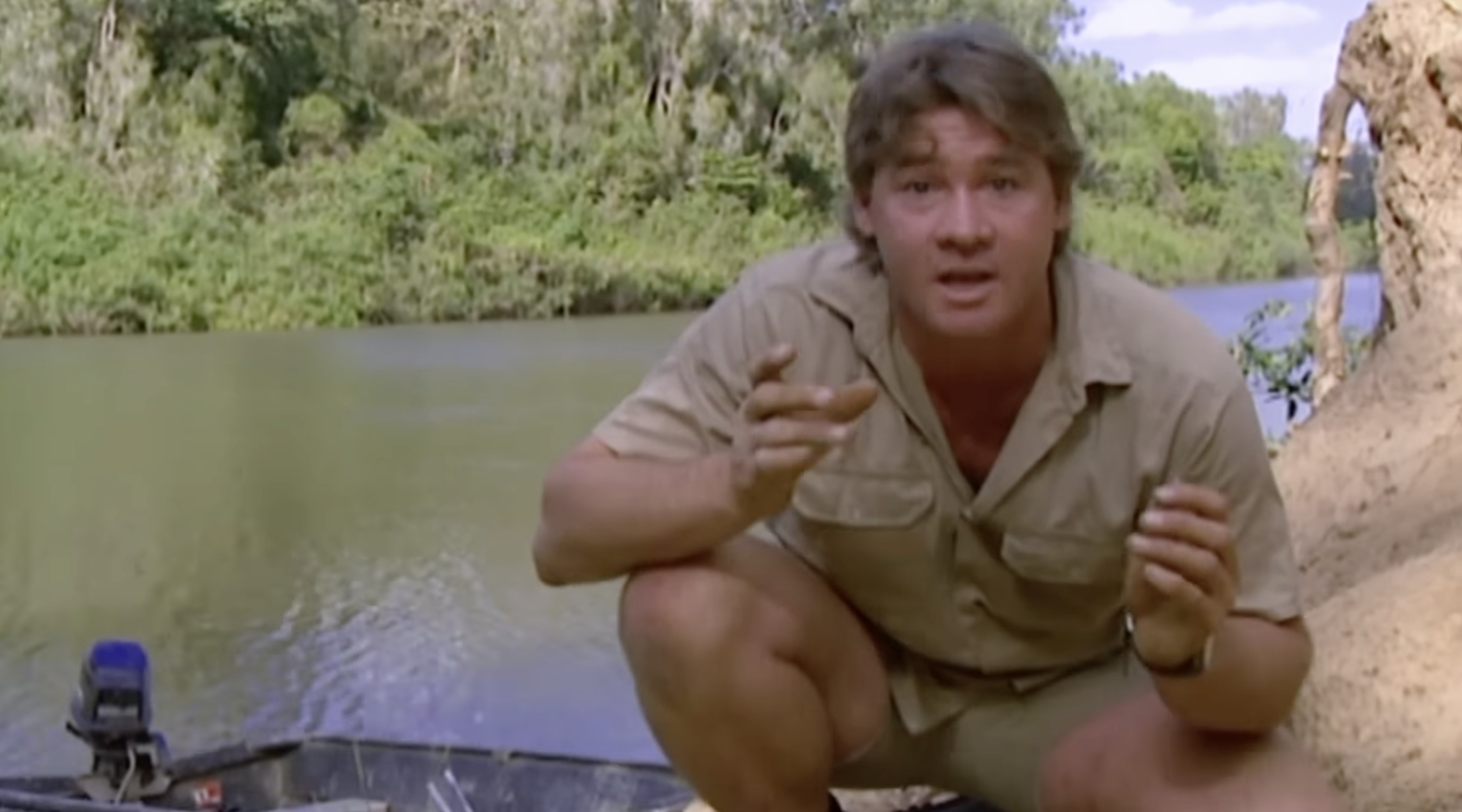 Person crouching near a riverbank, gesturing with hands, in a natural setting with trees and water in the background
