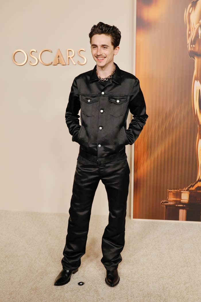 Person in sleek black suit on a red carpet, hands in pockets, Oscars logo visible in background