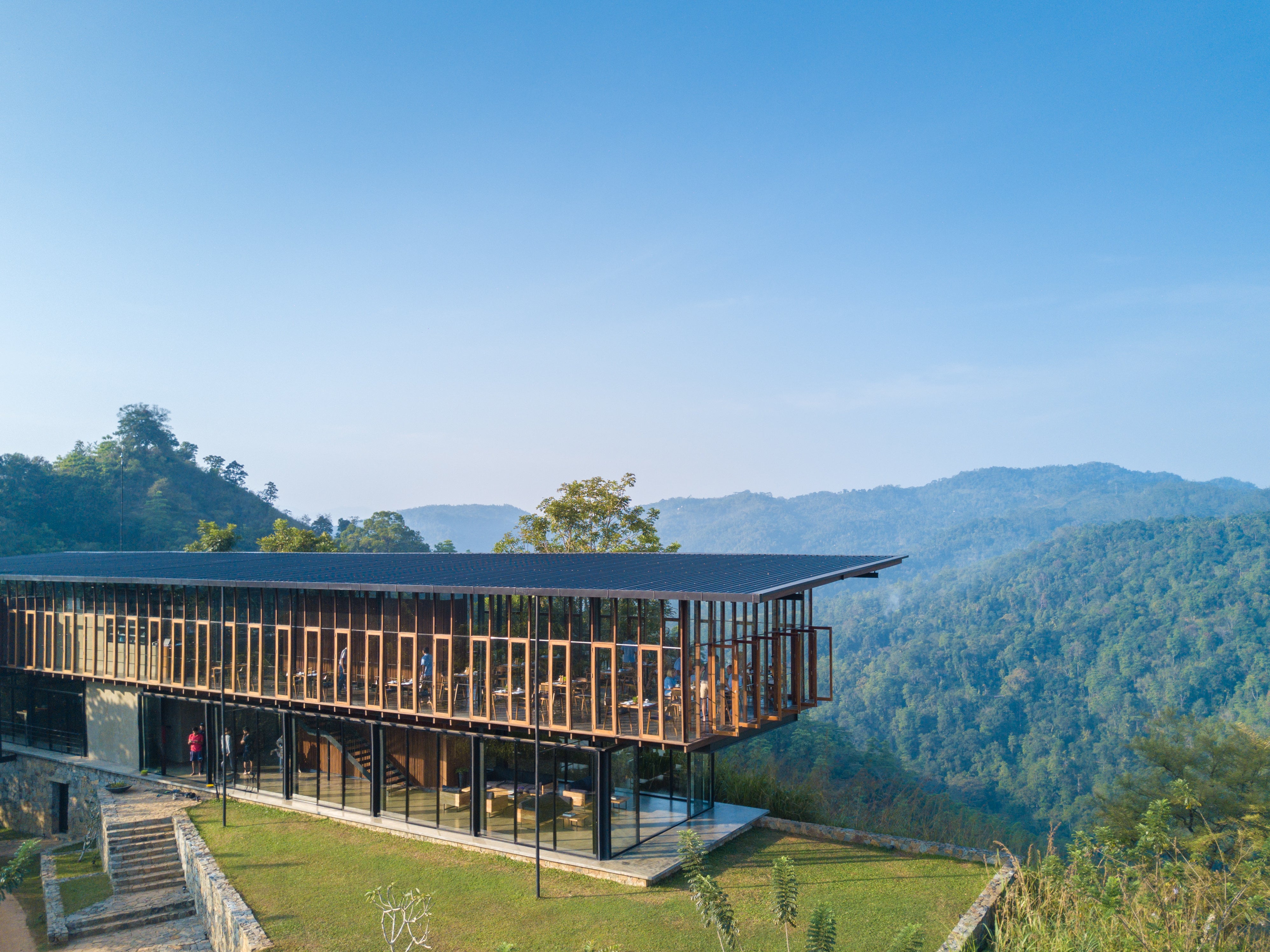 Modern glass and wood building on a hillside overlooking lush mountains under a clear sky