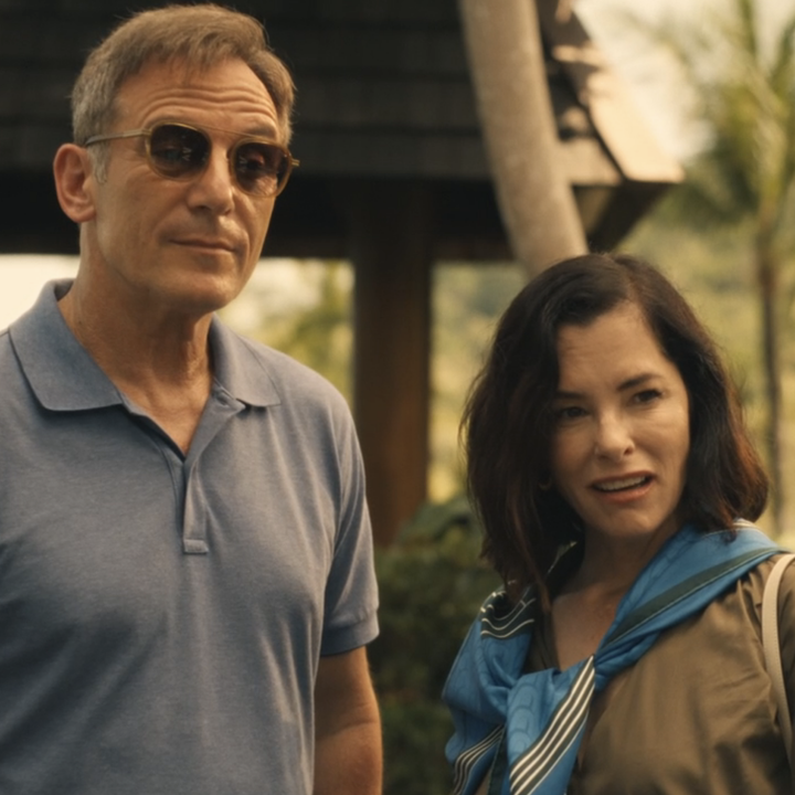 Two individuals stand outdoors, both wearing casual clothing. The man has on sunglasses and a polo shirt, while the woman wears a jacket