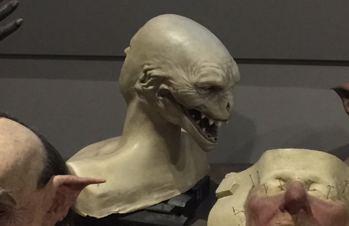 Realistic sculpted bust of a creature with pointed ears, sharp teeth, a wrinkled face, a wide, stretched mouth, and a smooth, elongated head, displayed in an art studio setting
