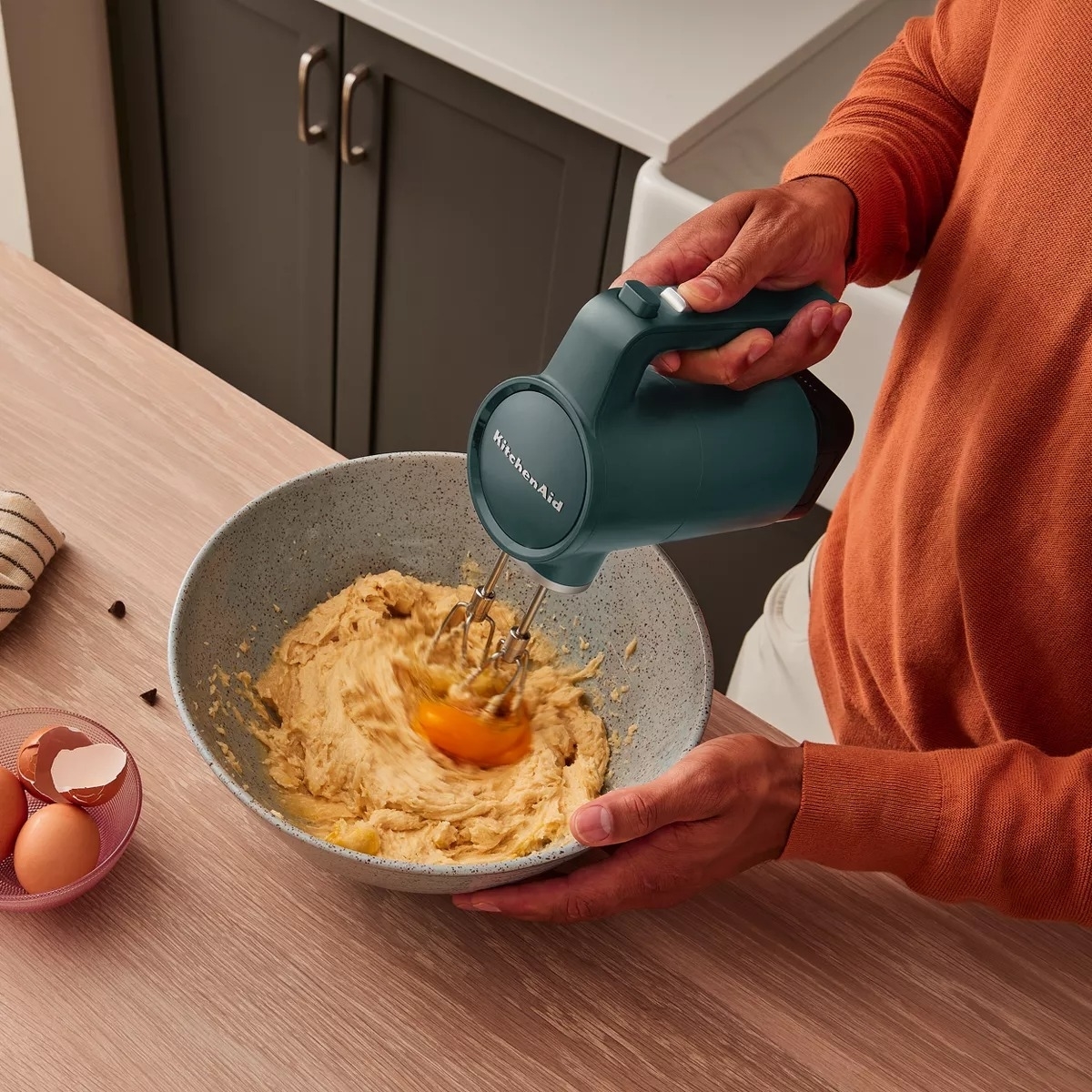 Person using a KitchenAid hand mixer to combine ingredients in a bowl on a kitchen counter