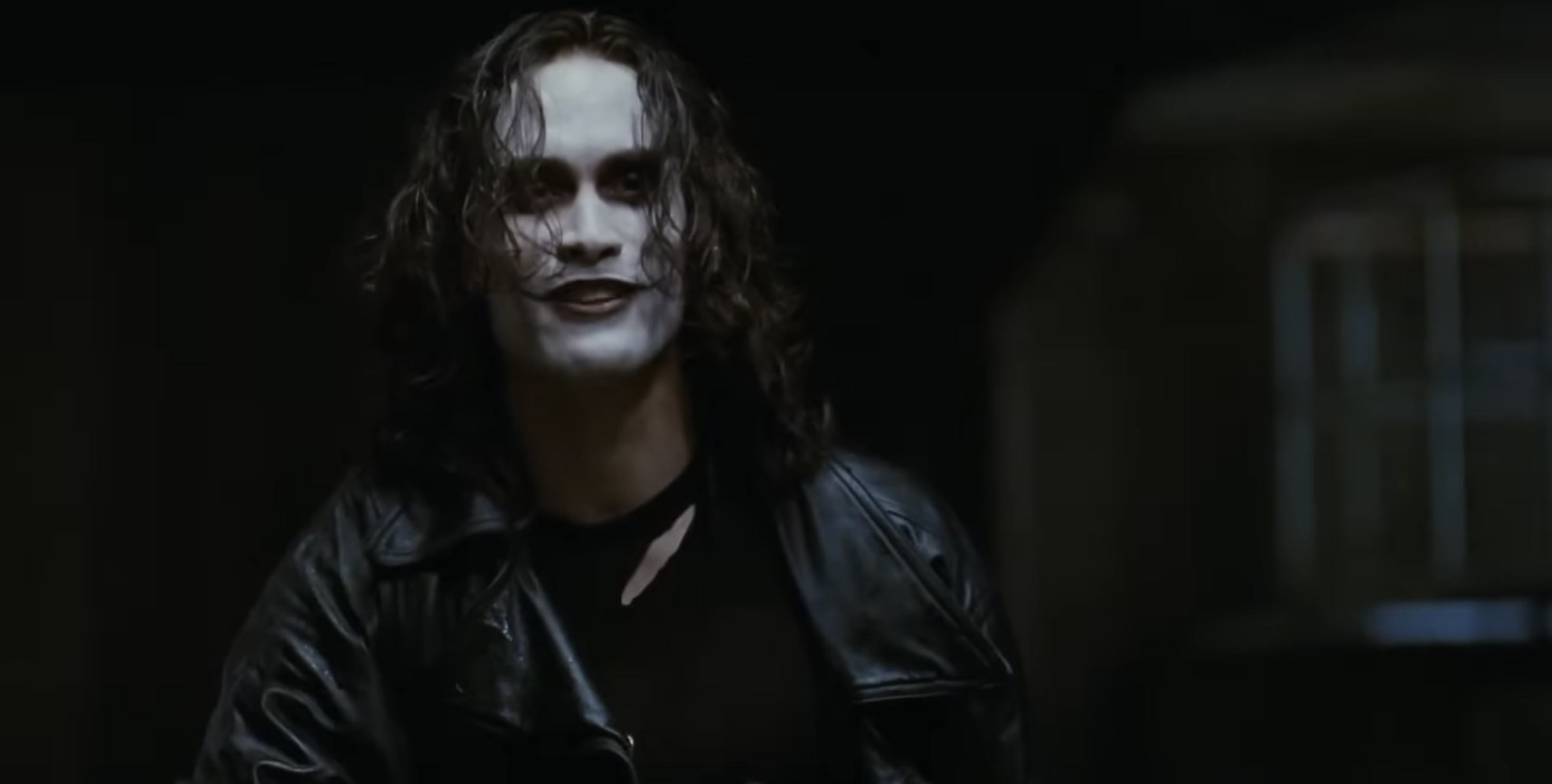 An actor with long hair in a dark leather jacket, wearing theatrical makeup, appears in a dramatic scene