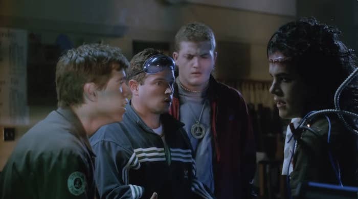 Group of four people having an intense conversation indoors at night, with one person wearing futuristic cables and equipment