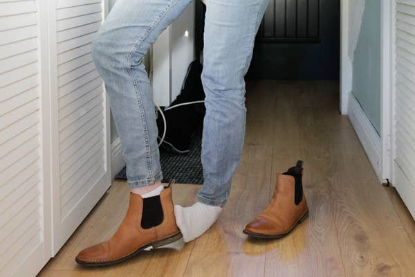Person wearing jeans stepping out of a brown ankle boot with one foot, while wearing a white sock, in a hallway