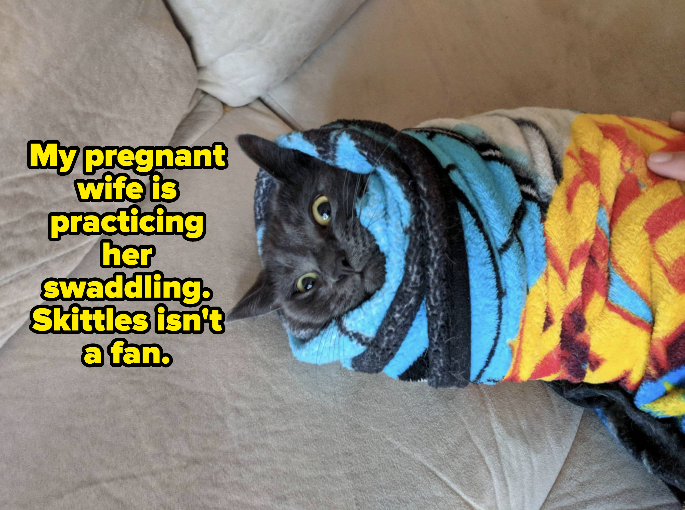 A cat is wrapped snugly in a colorful blanket, with only its head peeking out. Its eyes are wide open, and it rests on a couch