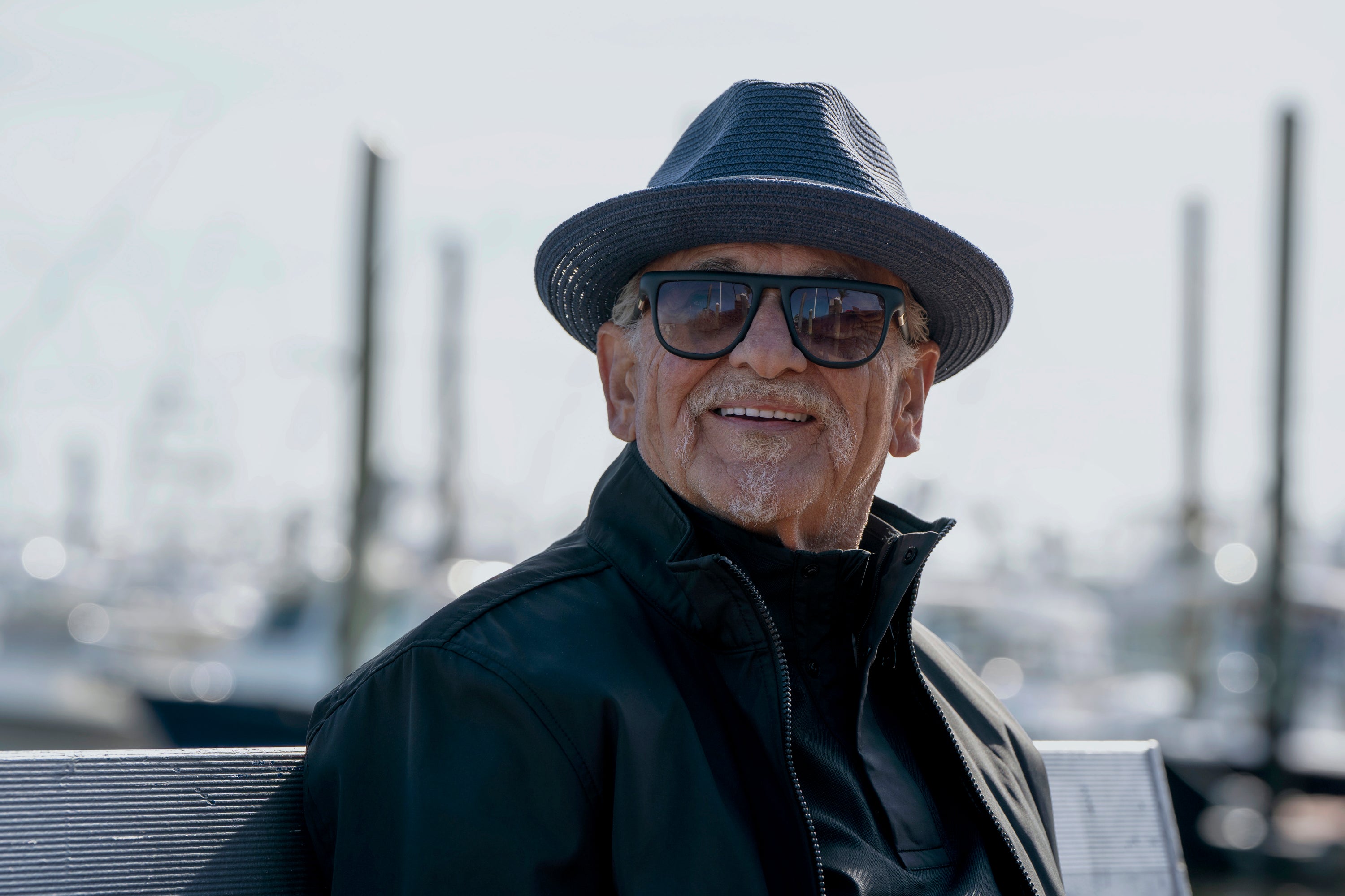 Person outdoors wearing a hat, sunglasses, and a dark jacket, smiling while sitting on a bench