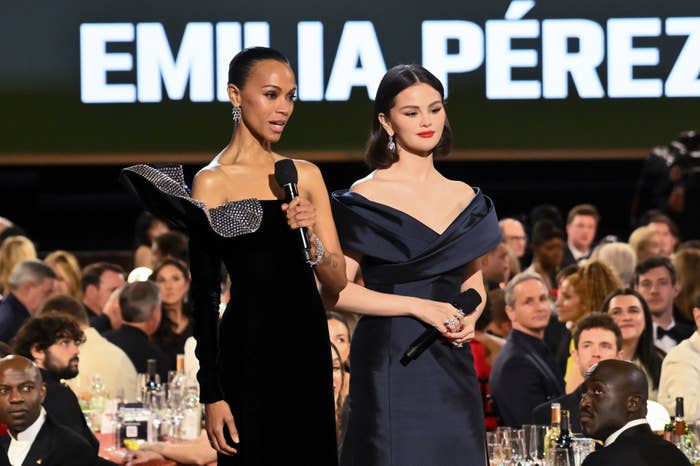 Two women in elegant evening gowns hold microphones on stage at a formal event, with a crowd seated behind them. Large text in the background reads "EMILIA PÉREZ."