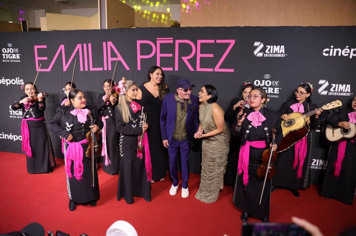 Group of people on a red carpet, including several musicians with string instruments and a person in a patterned dress, posed in front of a backdrop