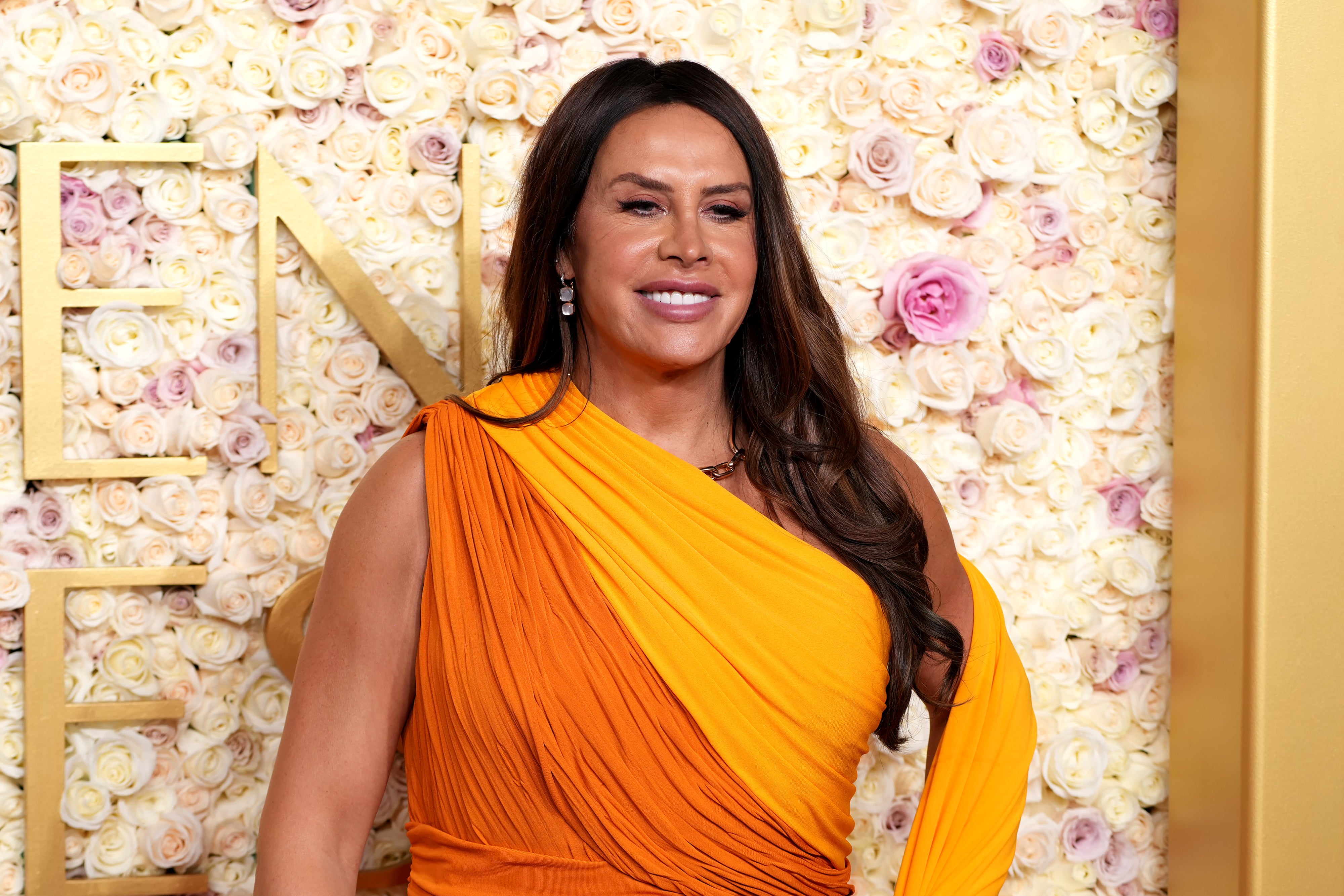 Person in elegant orange gown poses against a floral backdrop, smiling