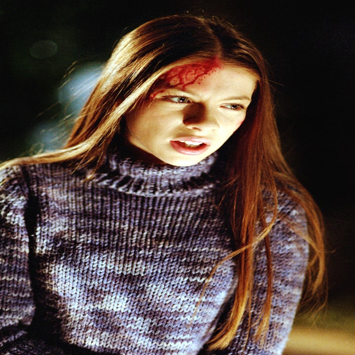 Person with long hair wearing a knit sweater has a makeup effect resembling a head wound, appearing in character for a dramatic scene