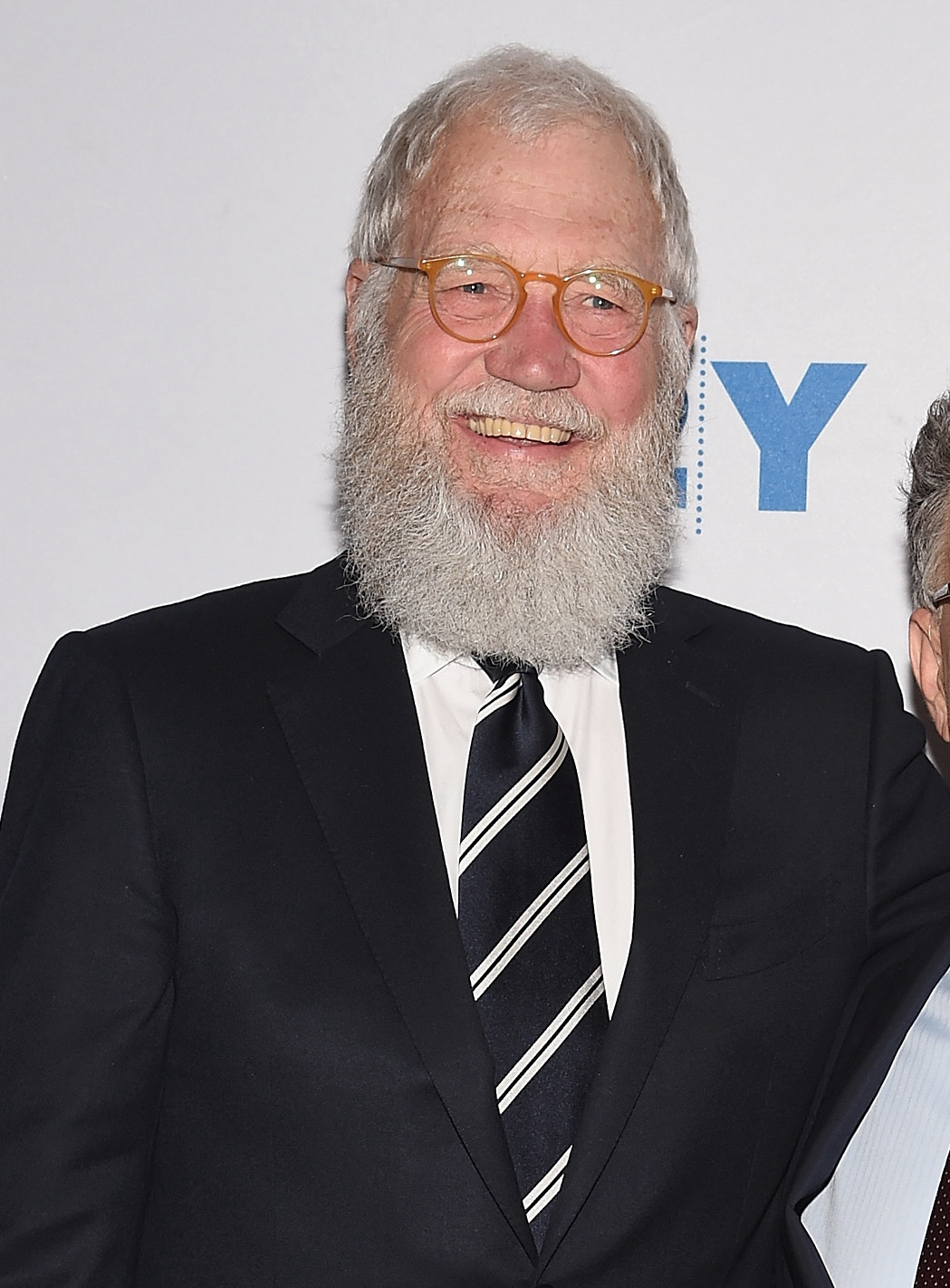 Man with a achromatic  beard and glasses successful  a achromatic  suit   and striped tie, smiling astatine  a personage  event