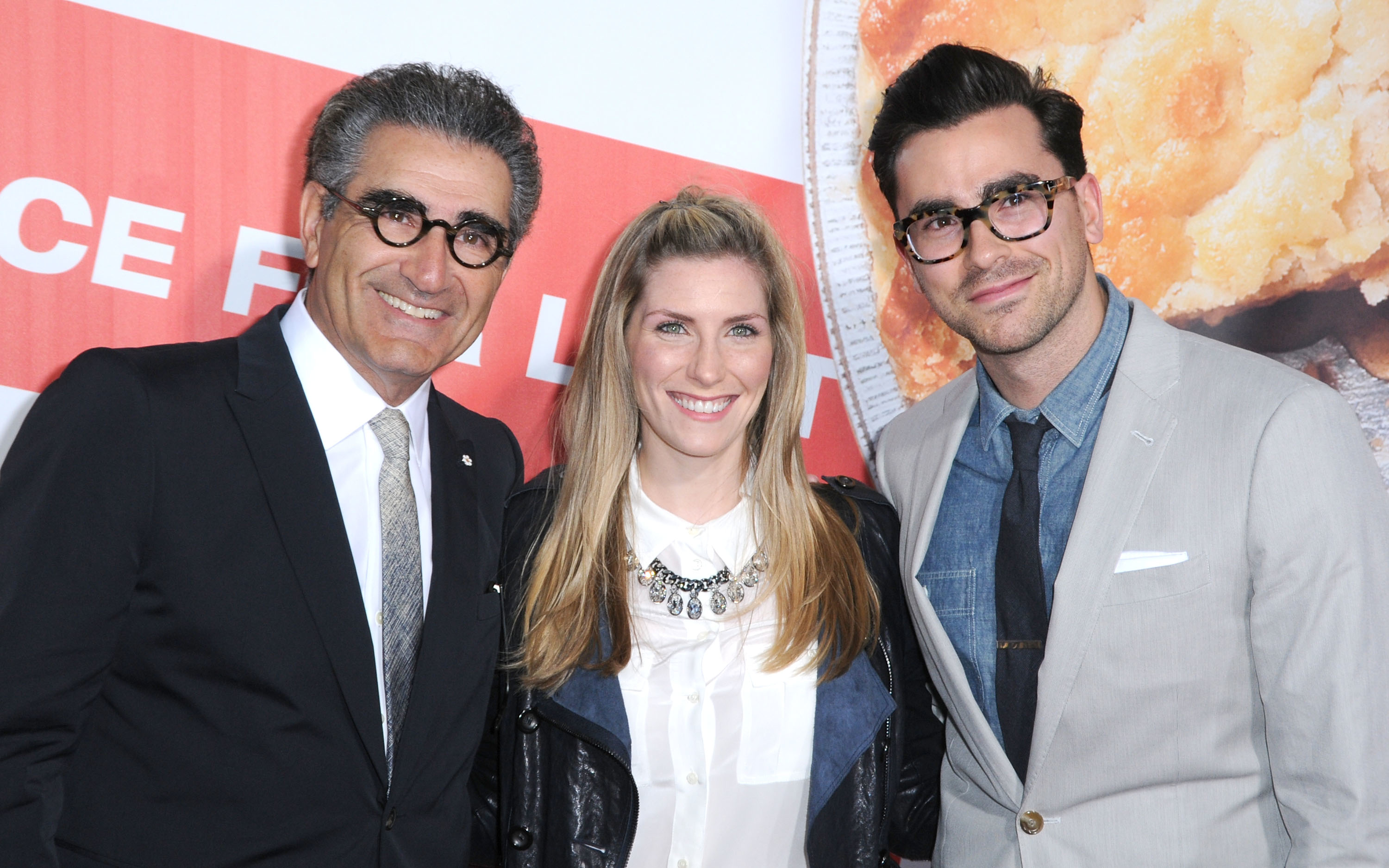 Three radical   smiling connected  a reddish  carpet; 2  men successful  suits and glasses, and a pistillate   successful  a blouse with a layered necklace