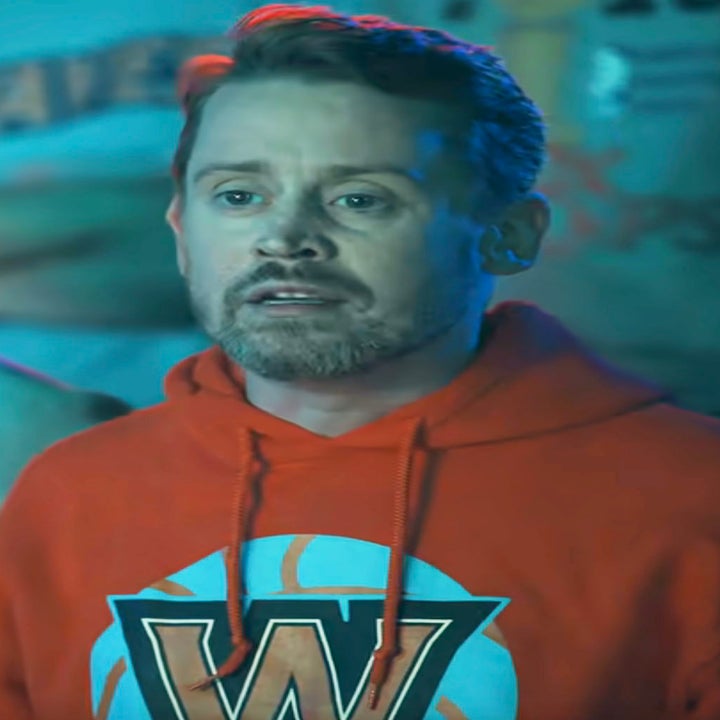 A man with facial hair wearing a hoodie with a "W" design, looking concerned, stands among others at an event