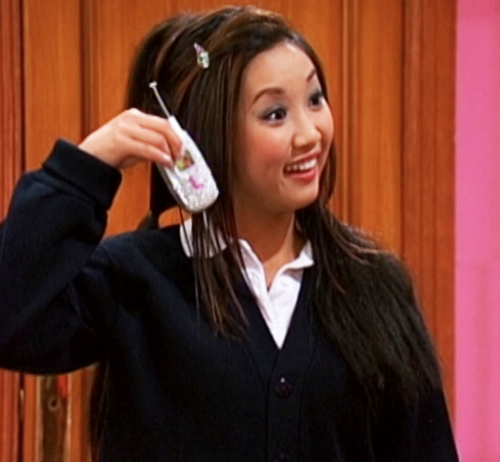 Person in a black cardigan holds a small flip phone near their ear, smiling enthusiastically in a room with wooden panels