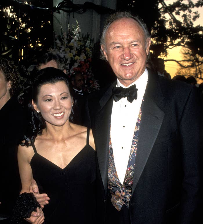 Gene Hackman în smoking cu vestă cu model stă alături de Betsy în rochie neagră elegantă la un eveniment. Ei zâmbesc pentru cameră