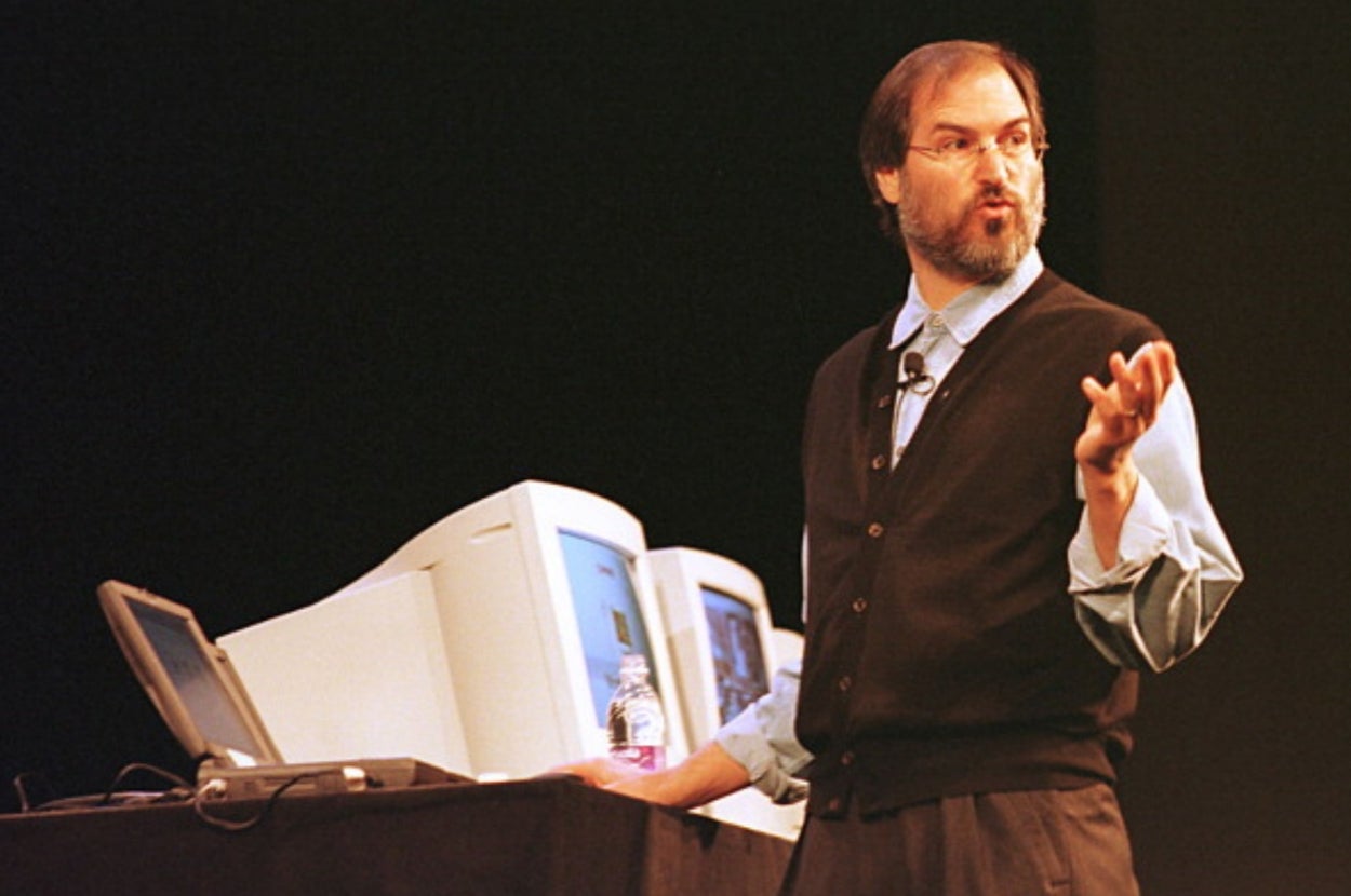 A person in a sweater vest and shirt is speaking passionately on stage beside vintage computer monitors and a laptop