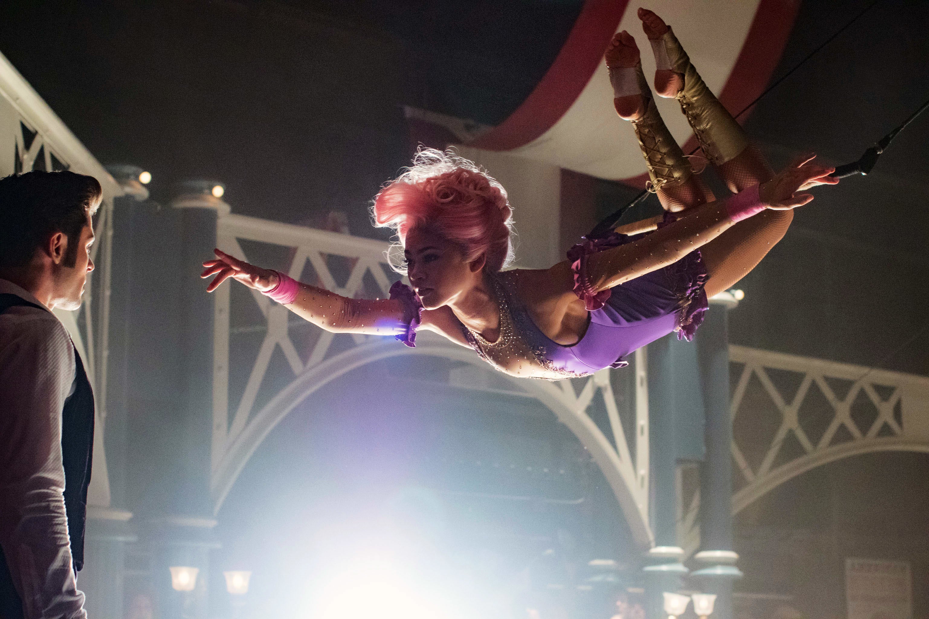 Aerial performer with styled hair in a leotard gracefully suspended mid-air during a circus act, observed by a person on the ground