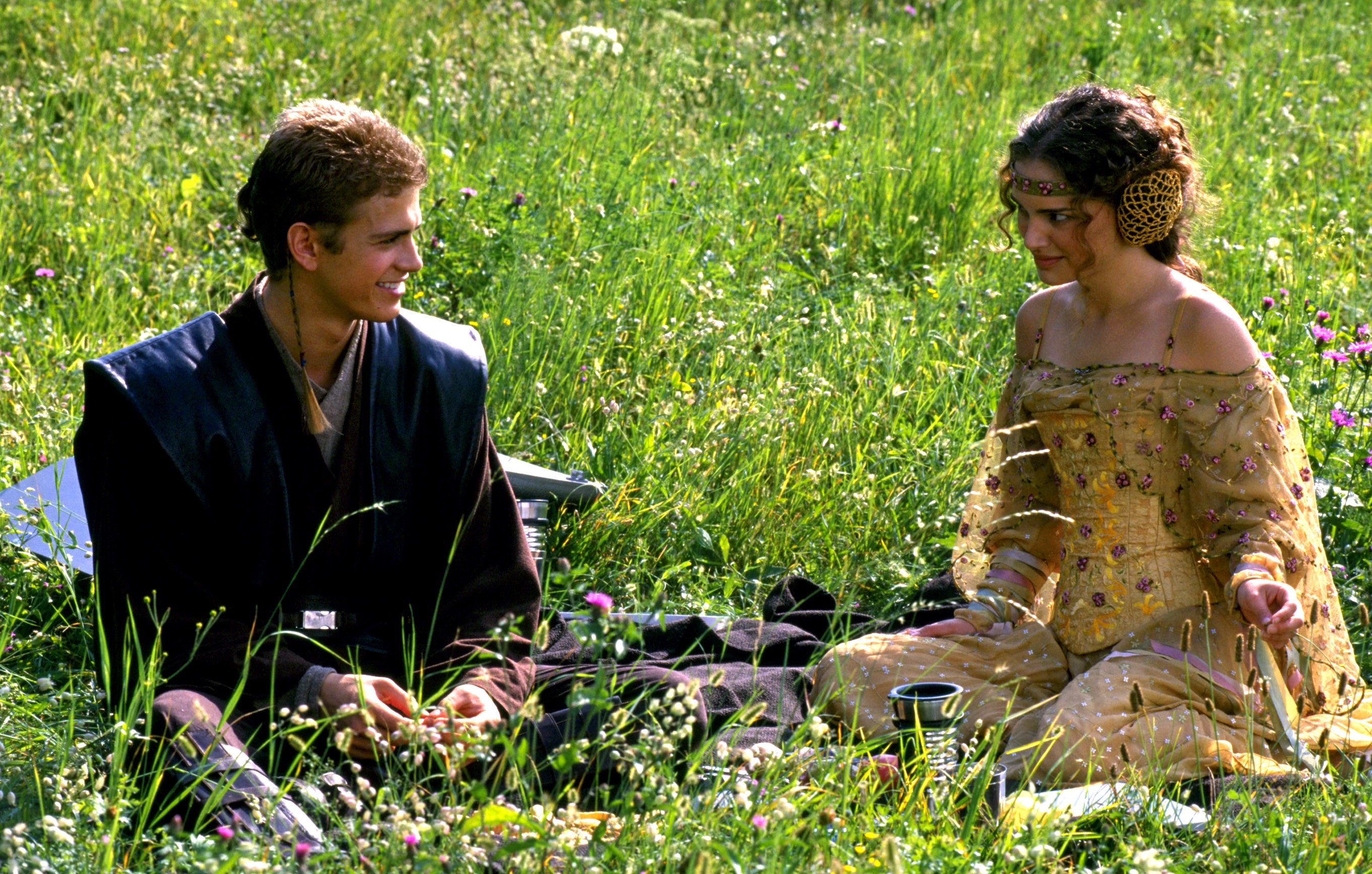 Two characters sit in a grassy field, dressed in elaborate, ornate costumes resembling a scene from a sci-fi or fantasy movie
