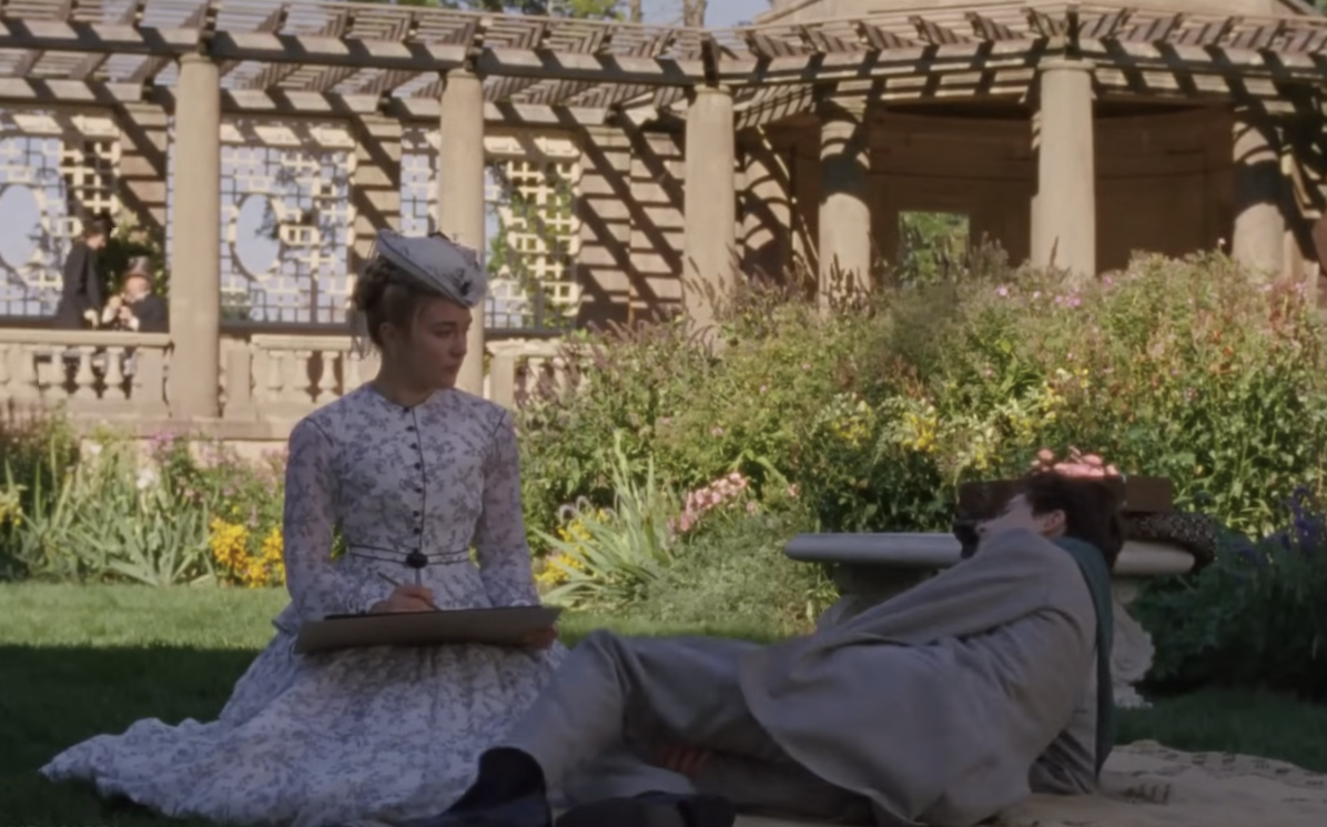 Scene from a period drama with a woman in Victorian attire sketching outdoors near a lounging man in a garden setting