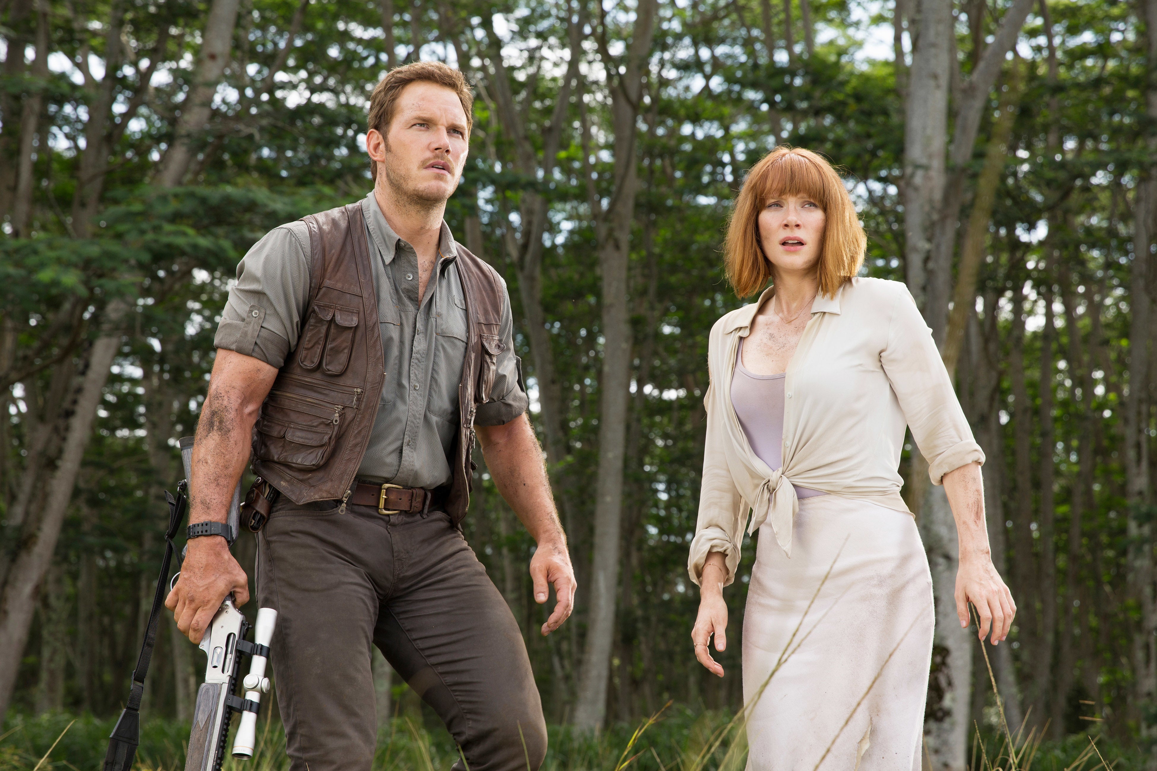 Two people in outdoor attire stand alert in a forest setting, appearing ready for action