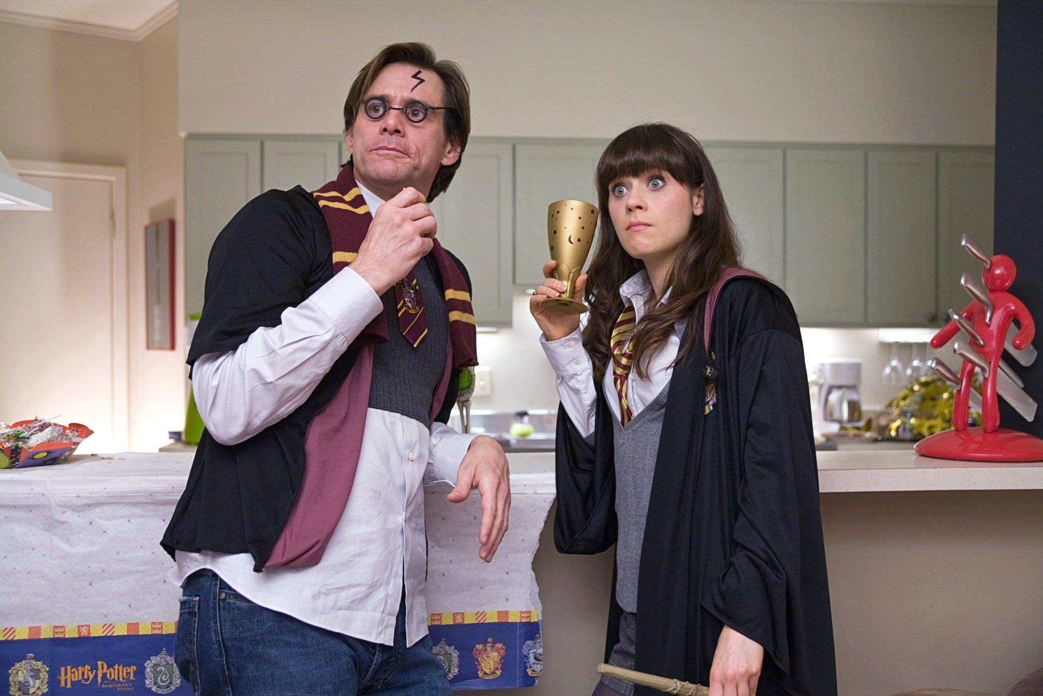 Two people in Hogwarts-themed costumes, holding a wand and goblet, in a kitchen. They're dressed as wizard students with robes and scarves