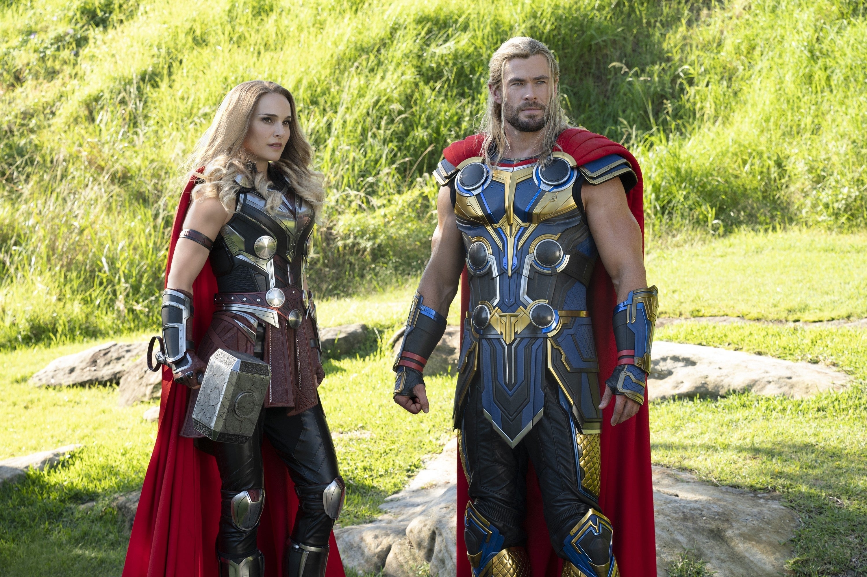 Two actors portray armored superheroes with red capes, standing on grass in a natural setting, resembling Norse mythology characters