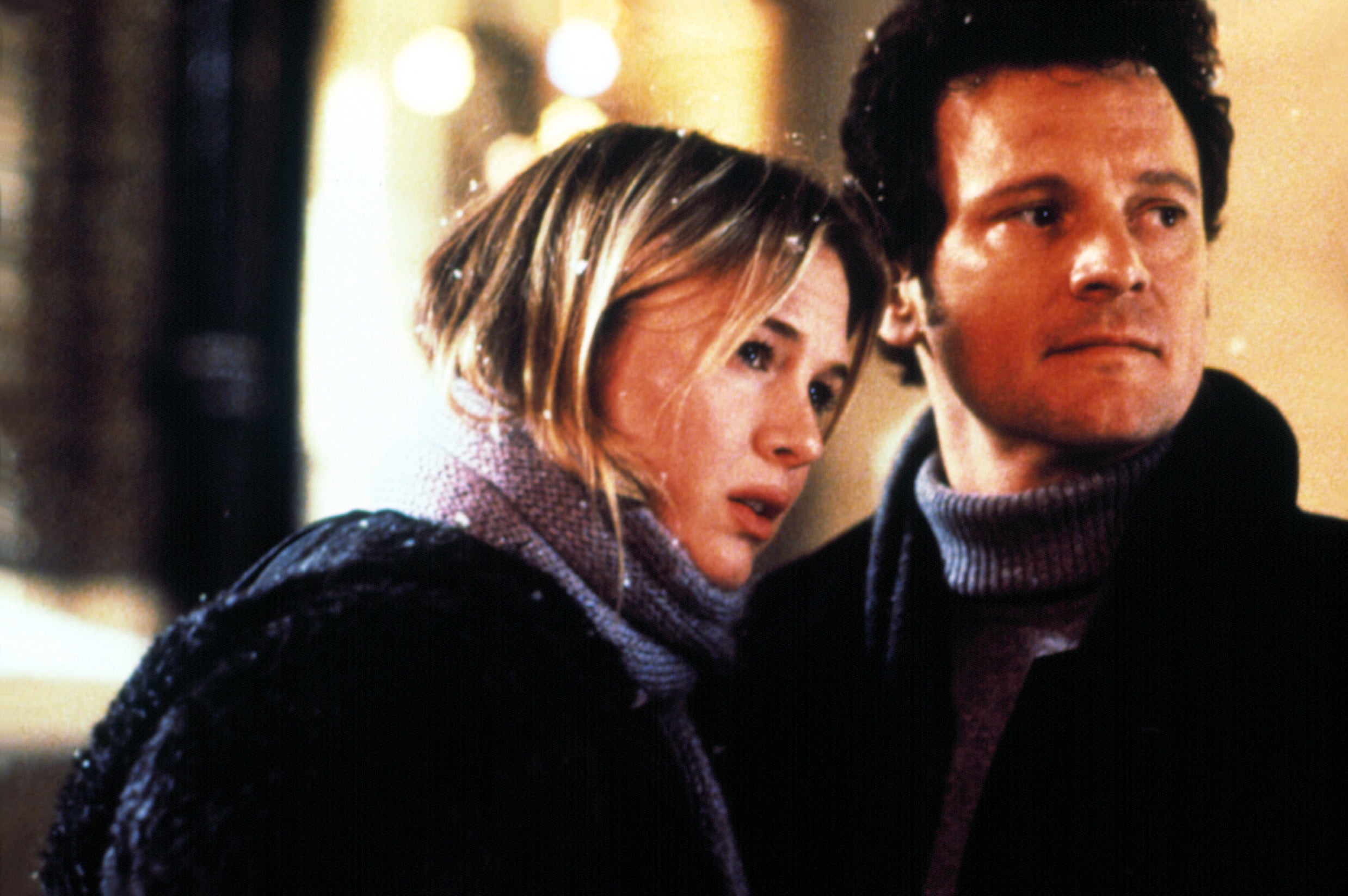 Two people stand closely together, wearing winter coats and scarves, looking forward with concerned expressions