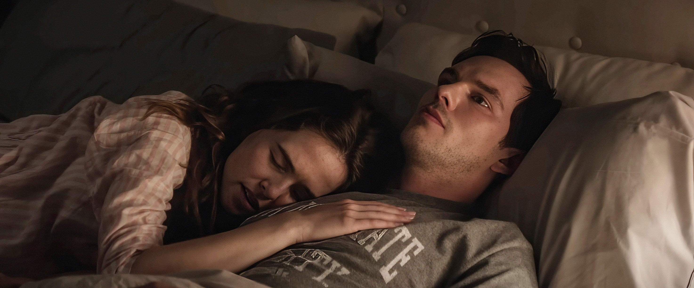 A couple lies in bed; one person rests their head on the other's chest, who looks awake and thoughtful