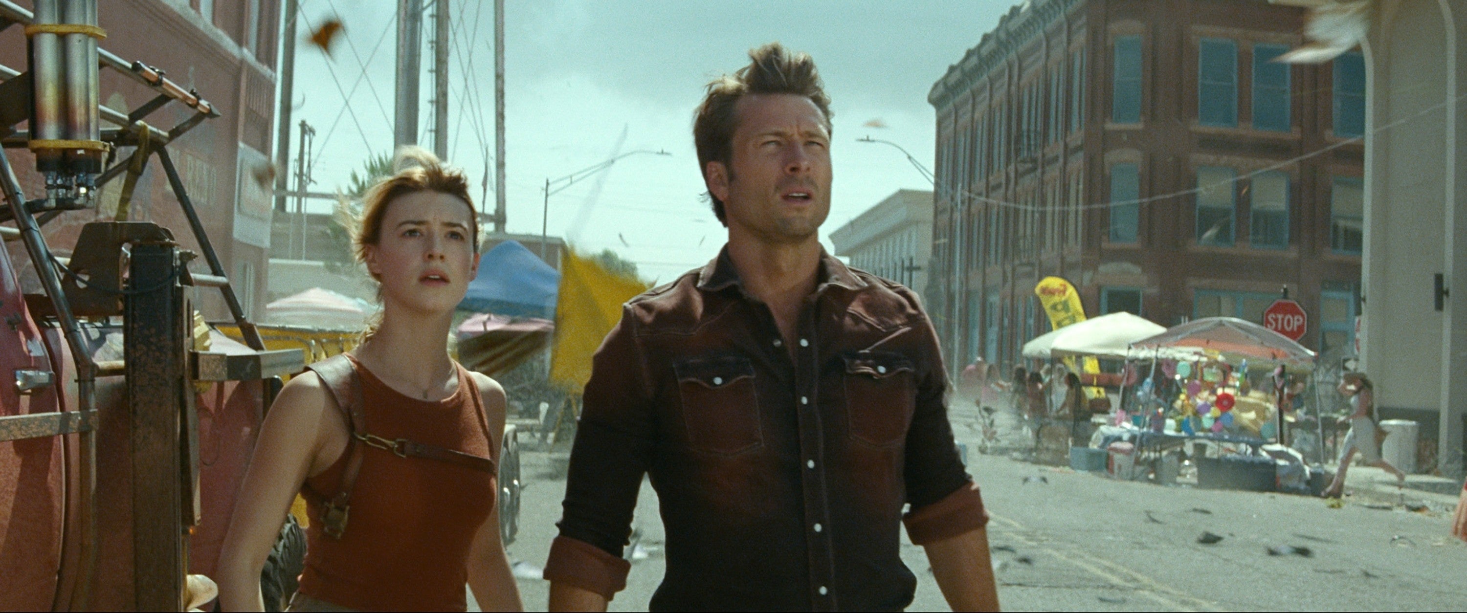 Two people look up with concern on a street filled with debris, stalls, and distant buildings