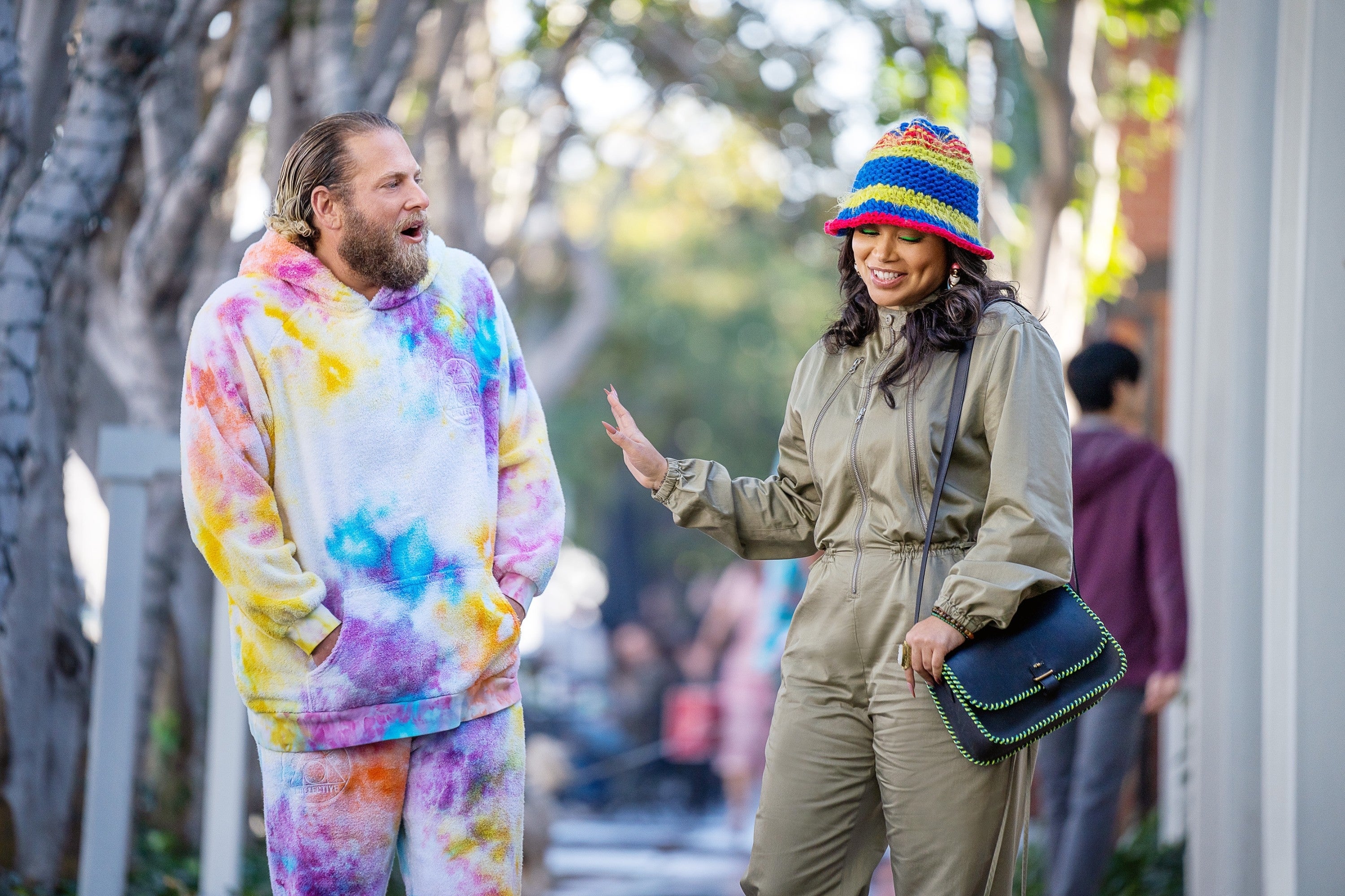 Two people outdoors; one in a vibrant tie-dye tracksuit, and the other in a stylish jumpsuit with a colorful crochet hat, in casual conversation