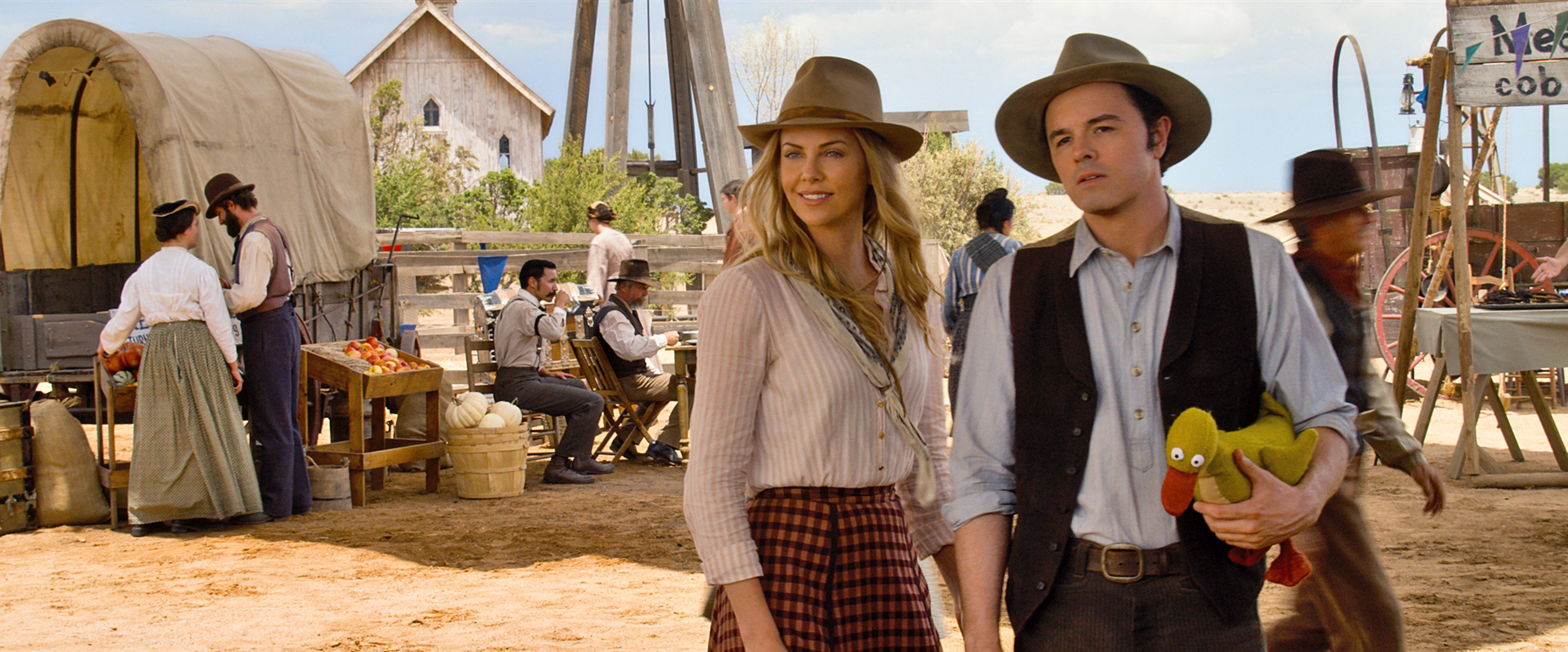 Two people dressed in Old West attire stand in a Western-style town setting. One holds a rubber chicken. Others in period costumes are in the background