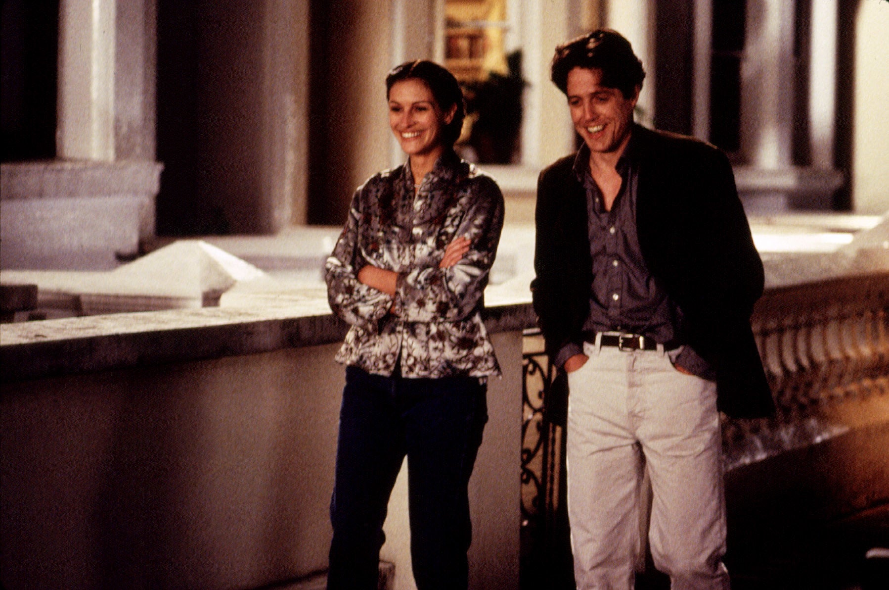 Two actors on a balcony, smiling. The woman wears a patterned blouse and jeans; the man wears a dark blazer with a shirt and light pants