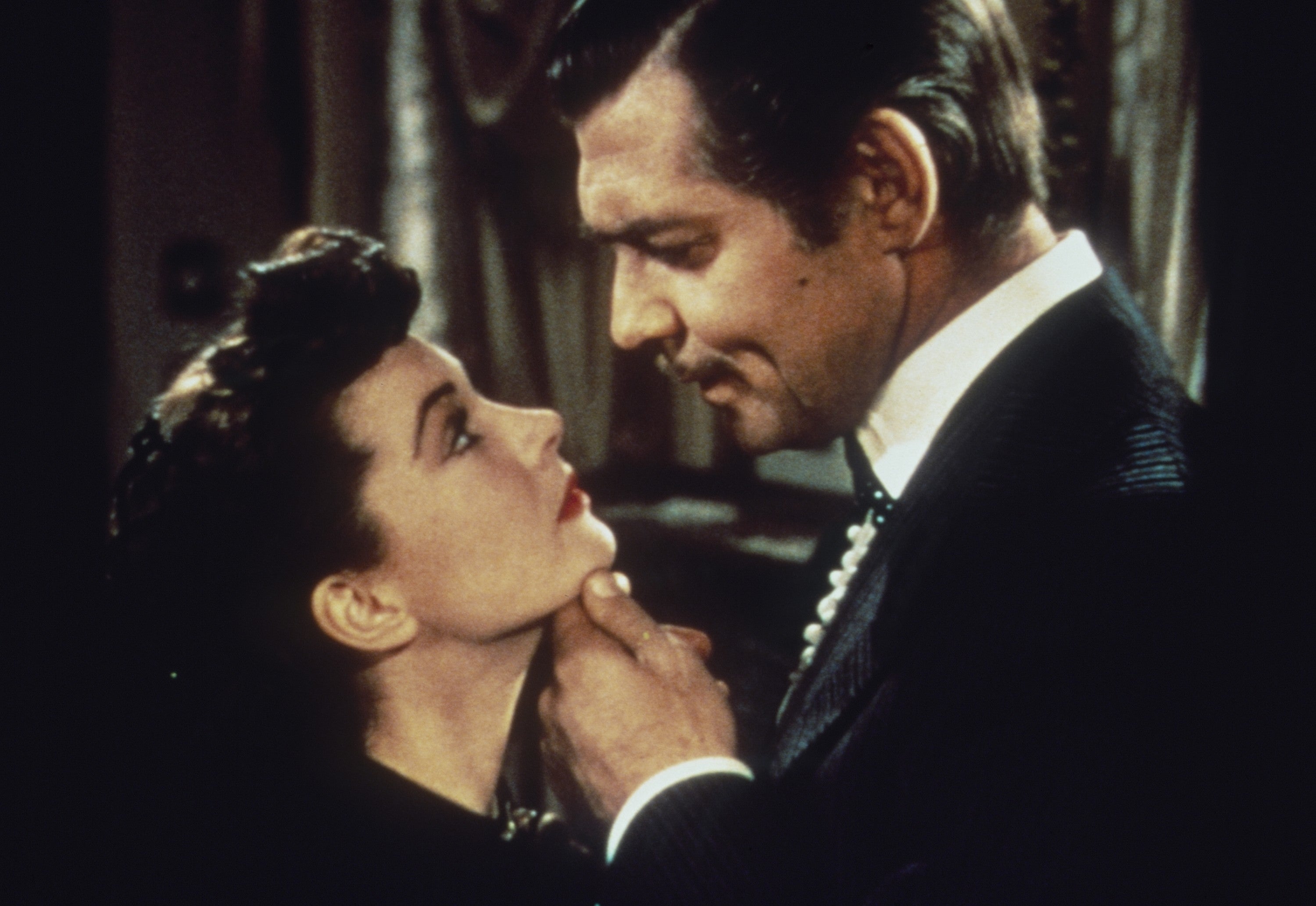 A man and woman gaze at each other intimately, both dressed in classic, elegant attire with formal hairstyles, embodying old Hollywood glamour