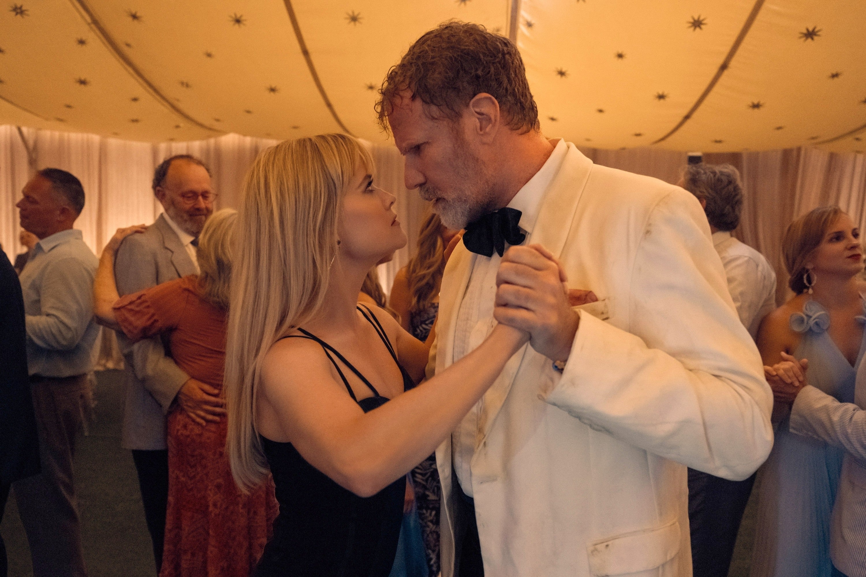 Man in white suit and bow tie dancing closely with woman in black dress at an elegant event under a decorated tent