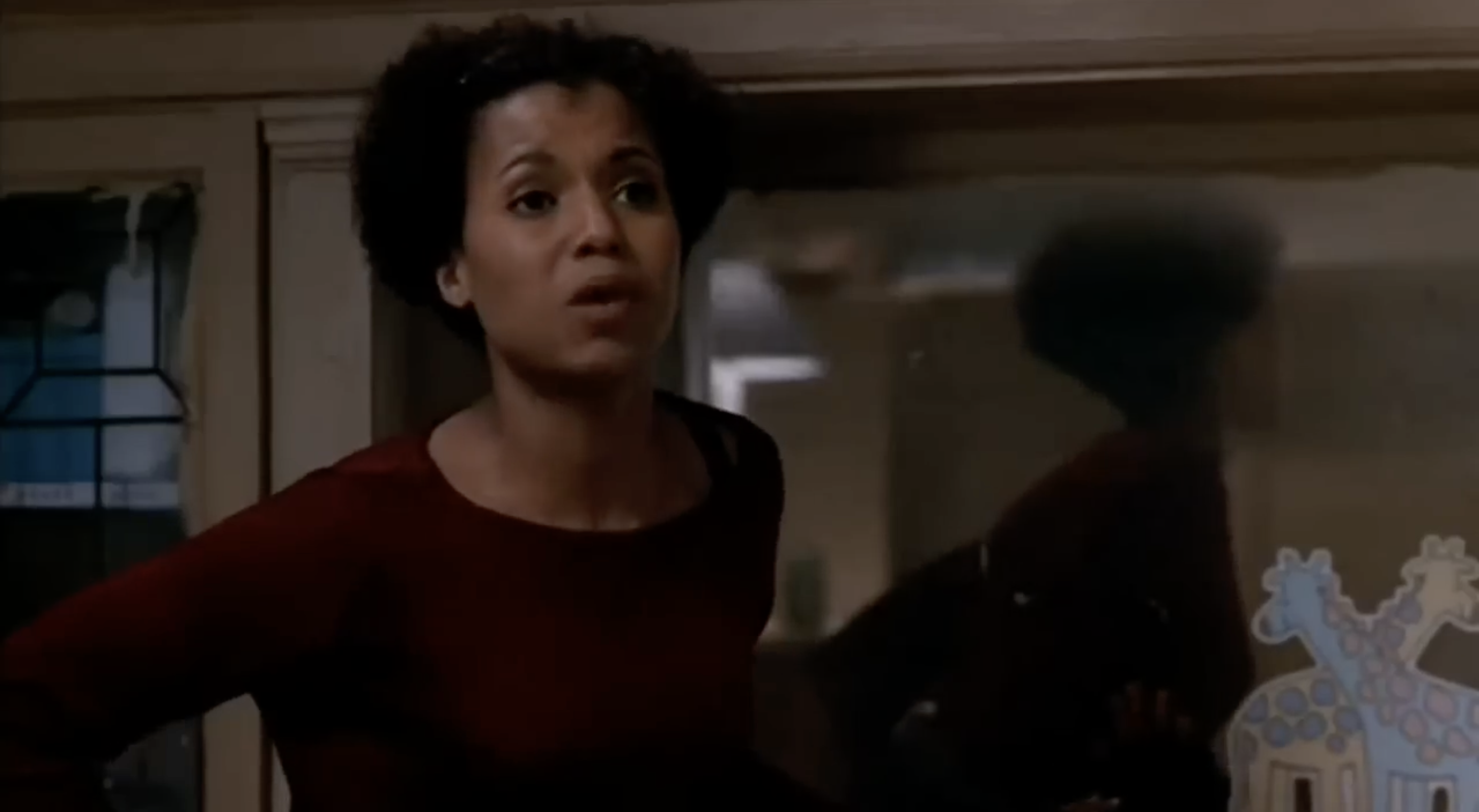 A person is indoors, speaking, in a living room setting. A giraffe decoration is visible on a window behind them