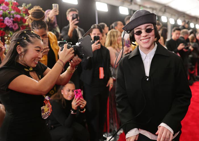 Person on red carpet wearing a stylish hat, black jacket, and sunglasses, smiling as someone takes their photo with a vintage camera