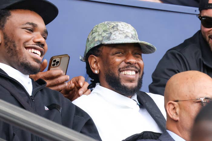 Two individuals smile while seated at an event. One wears a camo-patterned cap and a white shirt, the other a black jacket with a white shirt