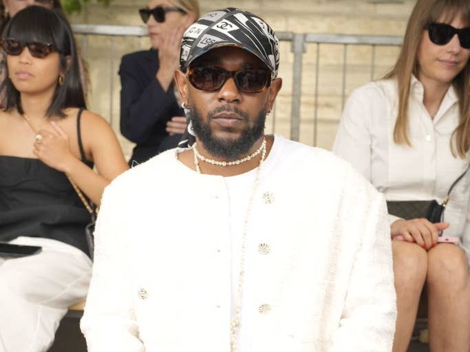 Person in a white textured jacket and patterned pants at a fashion event, sitting front row with people in sunglasses behind