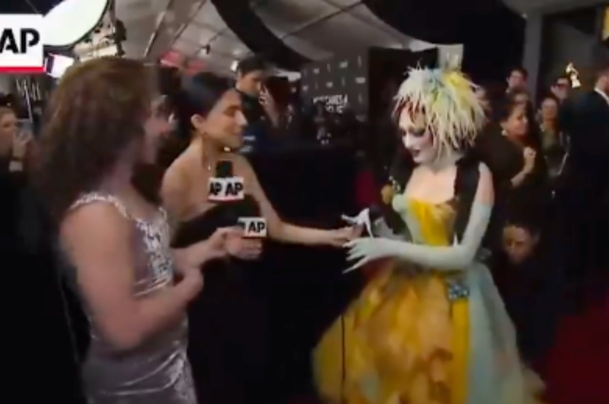 Two interviewers speak with a person in an extravagant, theatrical gown on the red carpet at a celebrity event