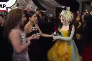 Two interviewers speak with a person in an extravagant, theatrical gown on the red carpet at a celebrity event