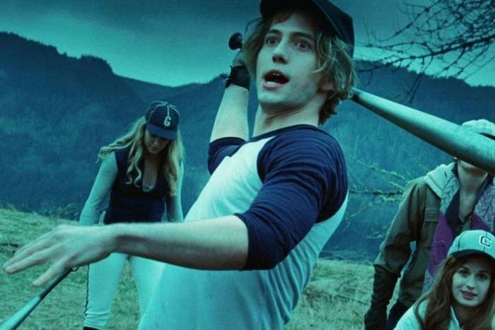 People playing baseball outdoors, wearing casual sportswear and caps. One person is swinging a bat mid-action, others watch in the background