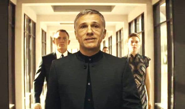 A man in a buttoned suit walks confidently down a corridor, followed by two others dressed formally