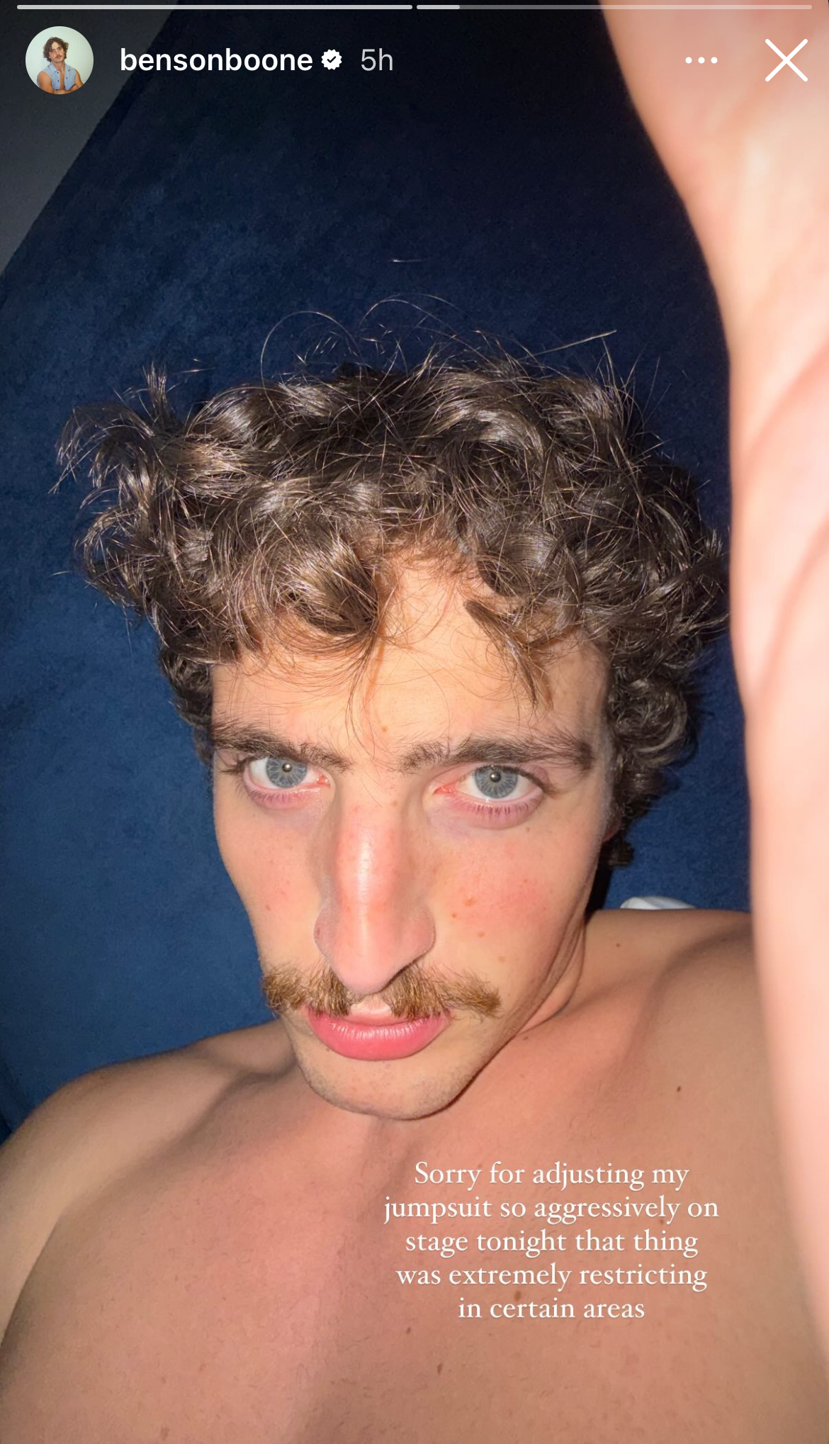 Benson Boone lying down with curly hair and a light mustache, looking at the camera. Text reads: "Sorry for adjusting my jumpsuit so aggressively on stage tonight; that thing was extremely restricting in certain areas"