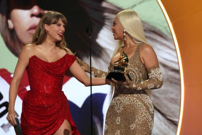 Two women on stage at an award show. One wears a red off-shoulder gown, the other a patterned dress, holding a Grammy. They're smiling and embracing
