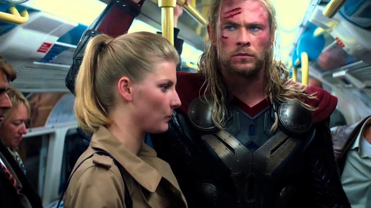 A person dressed as a superhero with armor and a cape stands beside another person in a busy train carriage